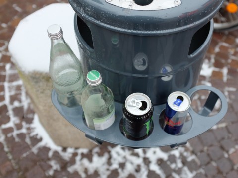 Grauer Müllbehälter mit Pfandring, in dem Flaschen und Dosen stecken.
